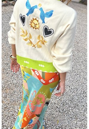 Person iført cremefarvet sweater med broderede blå fugle, sol, hjerter og grøn kant, kombineret med en farverig mønstret nederdel, stående på grus.