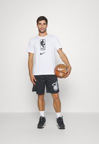 Fehér Nike póló NBA logóval, fekete Nike sort Miami Heat logóval, és fekete sportcipő, tart a kezében egy narancssárga kosárlabdát.