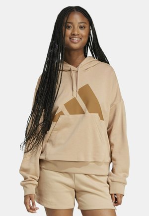 Giovane donna con lunghe treccine che indossa una felpa beige con un grande logo marrone e pantaloni corti beige abbinati, che sorride alla macchina fotografica.
