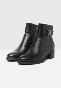 Bottines en cuir noir avec un bout rond, fermeture éclair et sangle décorative avec une boucle métallique. Talon bas en bloc. Texture lisse.