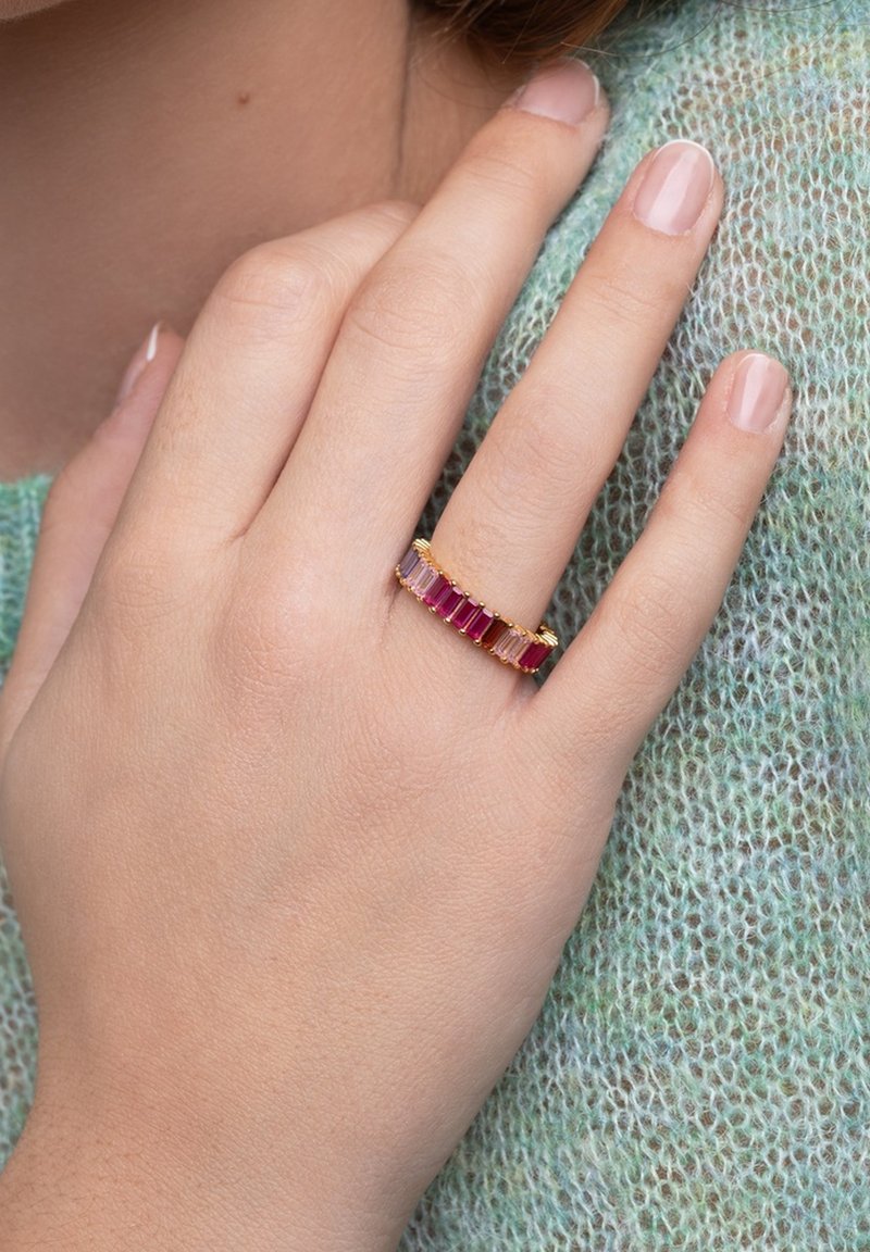 Main avec du vernis à ongles rose pâle portant une bague en or avec des pierres rectangulaires alternées roses et rouges sur un tissu en tricot vert.