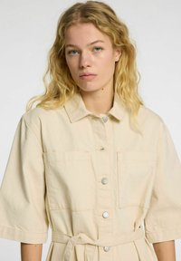 Beige bomulds skjortekjole med korte ærmer, der har krave, frontknapper, to brystlommer og et bindebælte i taljen.