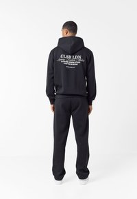 Άνδρας με την πλάτη, φοράει μαύρο φούτερ με κουκούλα με λευκό κείμενο «CLSR LDN Luxury British Atelier» και αντίστοιχο μαύρο παντελόνι με λευκά αθλητικά παπούτσια.