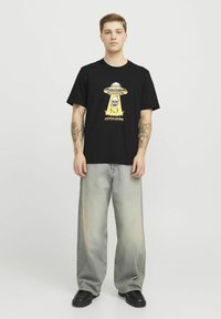 Sort bomulds t-shirt med et farverigt grafisk design af en UFO og kranie. Båret med lyse, løstsiddende jeans og sorte sko.