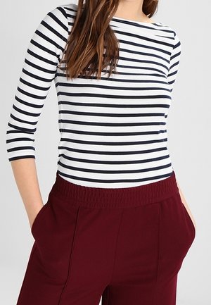 Žena oblečená v bílém a černém tričku s horizontálními pruhy a tříčtvrtečními rukávy, vysokými vínovými kalhotami s rukama v kapsách.