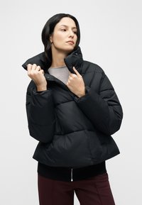Čierna nadýchaná bunda s vysokým golierom, prešívaným vzorom a elastickými manžetami. Materiál pôsobí hladko a leskle, so priliehavým strihom.