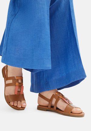 Brązowe, płaskie sandały z paskami i pomarańczowym akcentem, zestawione z niebieskimi, lekkimi spodniami o szerokich nogawkach. Gładka faktura, odkryte palce.