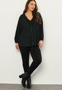 Blusa negra de tejido texturizado, con fruncido en la parte delantera, escote en V profundo y mangas largas tipo globo, combinada con pantalones negros ajustados y botines.