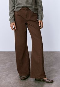 Pantalones de pierna ancha marrones combinados con un suéter gris y zapatos planos marrones oscuros con tachuelas, de pie sobre un suelo gris contra un fondo claro y liso.