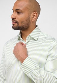 Uomo con barba che sistema il colletto di una camicia a righe leggera verde chiaro e bianca, rivolto a sinistra contro uno sfondo semplice.