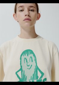 Mladá osoba s neutrálnym výrazom tváre nosí krémovú košeľu s potlačou zeleného usmievavého kresleného obrázku s dlhými vlasmi a rukami v bok.