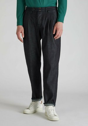 Pantalones de mezclilla oscura con un corte relajado, que cuentan con un frente plisado y puños doblados. Combinados con zapatillas blancas y una camisa verde de manga larga.