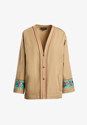 Veste beige avec un col en V, des boutons dorés et des garnitures rouges. Présente des broderies colorées à motifs sur les poignets. Tissu de poids moyen.