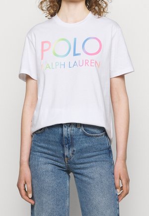 Osoba w białej koszulce z wielokolorowym napisem "POLO RALPH LAUREN" oraz niebieskich spodniach z wysokim stanem, pokazana od ramion do bioder.
