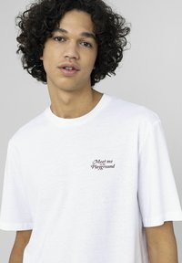 T-shirt en coton blanc à manches courtes avec un texte brodé en rouge "Rencontrez-moi sur le terrain de jeu" sur le côté gauche de la poitrine.