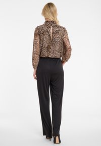 Woman with blonde hair in leopard print long-sleeve blouse and high-waisted black wide-leg pants, standing with hand on hip, facing away.