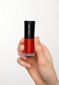 Płynna pomadka Lancôme w przezroczystej szklanej butelce z matowym, czarnym korkiem. Głęboki czerwony kolor, zaokrąglony kształt i złoty detal z logo.