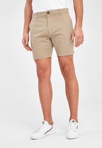 Beige korte broek van katoenmix, met een platte voorkant, riemlussen en een enkele knoopsluiting. Gemaakt in combinatie met witte sneakers.