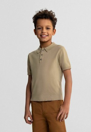 Jeune garçon aux cheveux bouclés souriant, portant un polo beige à manches courtes et un pantalon marron, debout devant un fond uni.