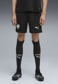 Vīrietis, kurš valkā melnas FC St. Pauli futbola šortus, melnas augstas ceļu zeķes ar "St. Pauli" un Puma logo, kā arī melnus futbola apavus uz pelēka fona.