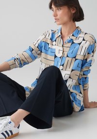 Camicia button-up in un tessuto a fantasia con grande tipografia in blu, crema e nero su uno sfondo chiaro. Maniche lunghe, vestibilità comoda.