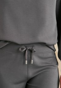 Szary komplet w postaci bluzy i dresów, wykonany z miękkiego, teksturowanego materiału, z regulowanym sznurkiem i metalowymi końcówkami na sznurku.
