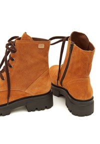 Botas de ante hasta el tobillo en marrón anaranjado con suelas de goma negra. Parte delantera con cordones y cremallera lateral. Presenta un pequeño logo en el costado exterior.