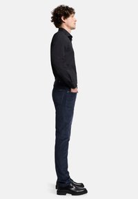 Camicia nera a maniche lunghe, design aderente, abbinata a jeans slim-fit blu scuro. Stivali neri con lacci completano l'outfit. Vista laterale.