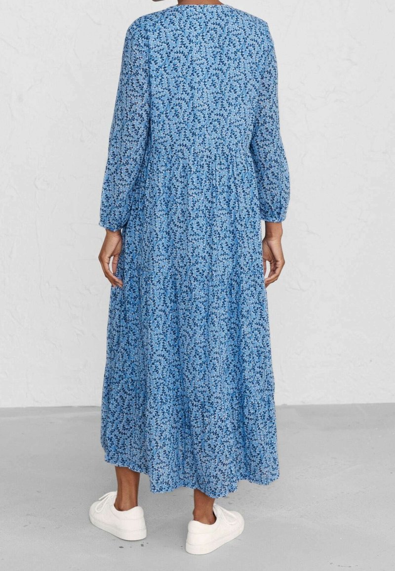 Blumenmaxikleid in Blau mit langen Ärmeln und geraffter Taille. Hat eine fließende Silhouette und dezente Blattmuster. Kombination mit weißen Sneakers.