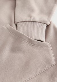 Hellbeiges Sweatshirtmaterial mit geripptem Bündchen und Tasche. Glatte Textur, präzise Nähte und klare Linien betonen das schlichte Design.