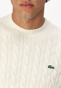 Cream cable-knit sweater featuring a ribbed crew neck and a small green crocodile logo on the chest. Soft texture with a classic design.
