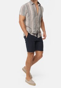 Kortærmet skjorte med knapper og lodrette striber i beige, brun og grøn. Kombineret med mørkeblå shorts og lyse espadrillos.