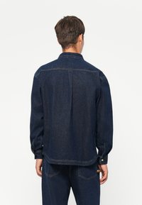 Dark blue denim shirt with long sleeves, button cuffs, pointed collar, and a straight cut. Features back yoke and side hem detailing.
