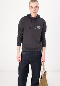 Felpa grigia in cotone con cappuccio e cordini, dettaglio del logo frontale, abbinata a jeans in denim scuro e una borsa a tracolla beige.