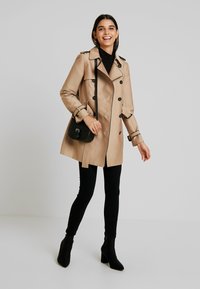 Beige trench coat with double-breasted buttons, shoulder epaulets, and a belt. Black skinny pants and ankle boots complement the outfit.