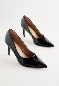 Zapatos de tacón alto de charol negro con punta afilada, una silueta elegante y un delgado tacón de aguja, con una textura suave y un interior nude.
