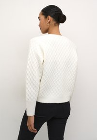 Pull blanc texturé avec un motif en losanges, des poignets et une ceinture côtelés. Porté avec un pantalon noir, vu de dos.