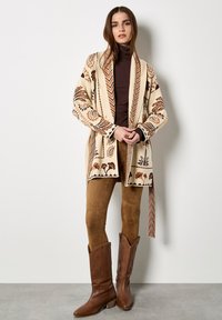 Sweter kardigan w kremowej bazie z misternymi brązowymi wzorami. Modelka łączy go z brązowymi zamszowymi legginsami i brązowymi wysokimi botkami.