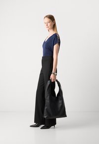 Schwarze Lederhandtasche mit weicher Textur, über der Schulter getragen. Model in eng anliegendem, dunkelblauem glitzerndem Top und weiten schwarzen Hosen, stehend vor hellem Hintergrund.