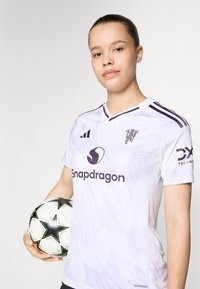 Bela nogometna majica z vijoličnimi detajli, ki ima v-izrez, tri črte na ramenih in detajle logotipa. Igralec drži žogo s staršnimi vzorci.
