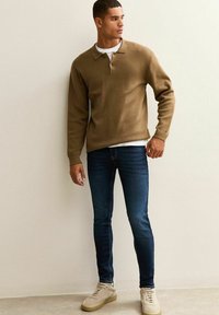 Brown knitted pullover with collar, paired with dark blue jeans and beige sneakers. Smooth texture with a sleek, fitted design.