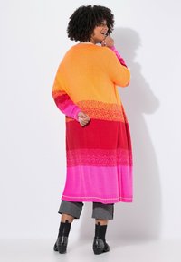 Donna con capelli ricci che indossa un lungo cardigan a strisce colorate nei toni dell'arancione, rosso e rosa, pantaloni grigi a tre quarti e stivali alla caviglia neri con borchie.