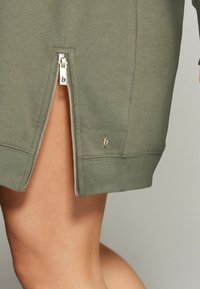 Olijfgroen sweatshirt met een zijrits, voorzien van ribgebreide manchetten en een onderrand, en een klein metalen logo nabij de rits. Gladde textuur.