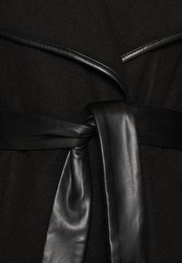 Svart ullplagg med ett slätt svart läderbälte knutet vid midjan. Tyget verkar mjukt, medan lädret tillför en kontrasterande textur.