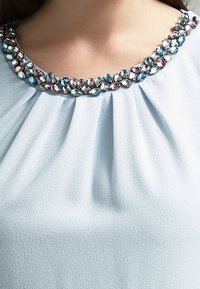 Blusa azul claro con pliegues en el escote, adornada con un cuello decorativo de gemas variadas en colores rosa, azul y transparente.