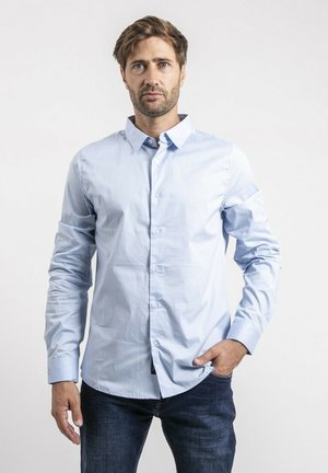 Homme aux cheveux bruns courts et barbe, portant une chemise bleue claire boutonnée et un jean foncé, debout avec une main dans la poche.