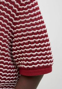 Vêtement en tricot texturé rouge avec des rayures blanches horizontales, des manches courtes et un ourlet côtelé aux manches. Le tissu semble superposé et confortable.