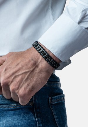 Black braided leather bracelet worn on wrist, set against a white shirt. Bracelet features a smooth texture and intricate weaving detail.