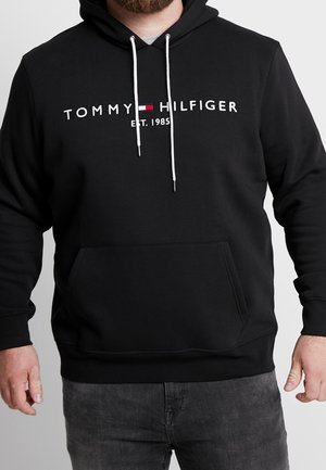 Mann trägt schwarzen Tommy Hilfiger Hoodie mit weißen Kordeln, Logo und dem Schriftzug "EST. 1985", kombiniert mit dunkelgrauen Jeans.