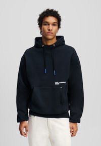 Crna hoodie izrađena od mješavine pamuka, s prednjim džepom u obliku kengura, kapuljačom s plavim detaljima, te logotipom otisnutim na prsima.